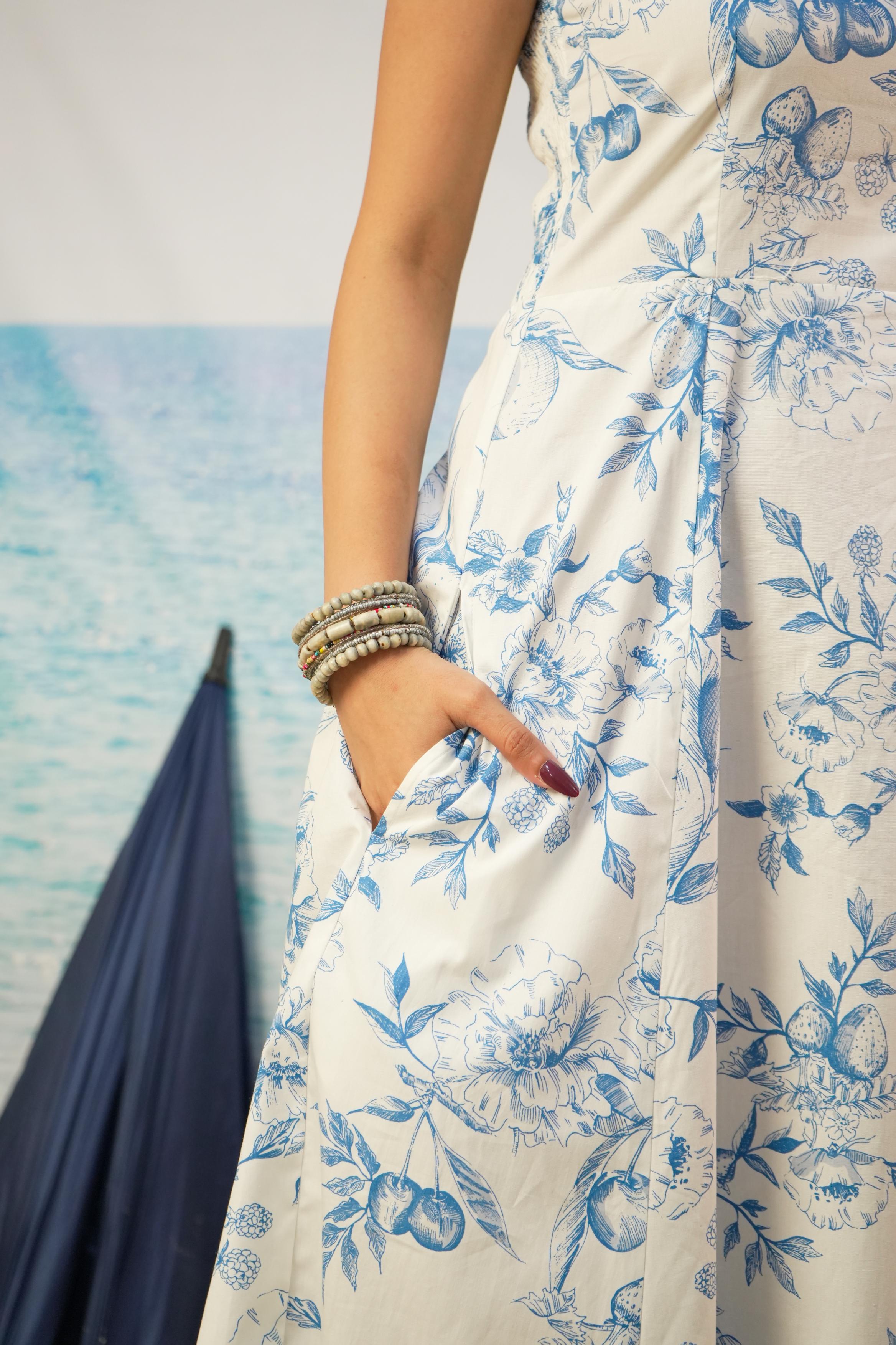 A close-up of a woman wearing a sleeveless white dress with a blue floral toile pattern, featuring pockets and a side slit, accessorized with stacked silver bracelets.