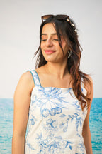 A woman with sunglasses on her head stands by the ocean wearing a white sleeveless top with a blue floral and fruit print.