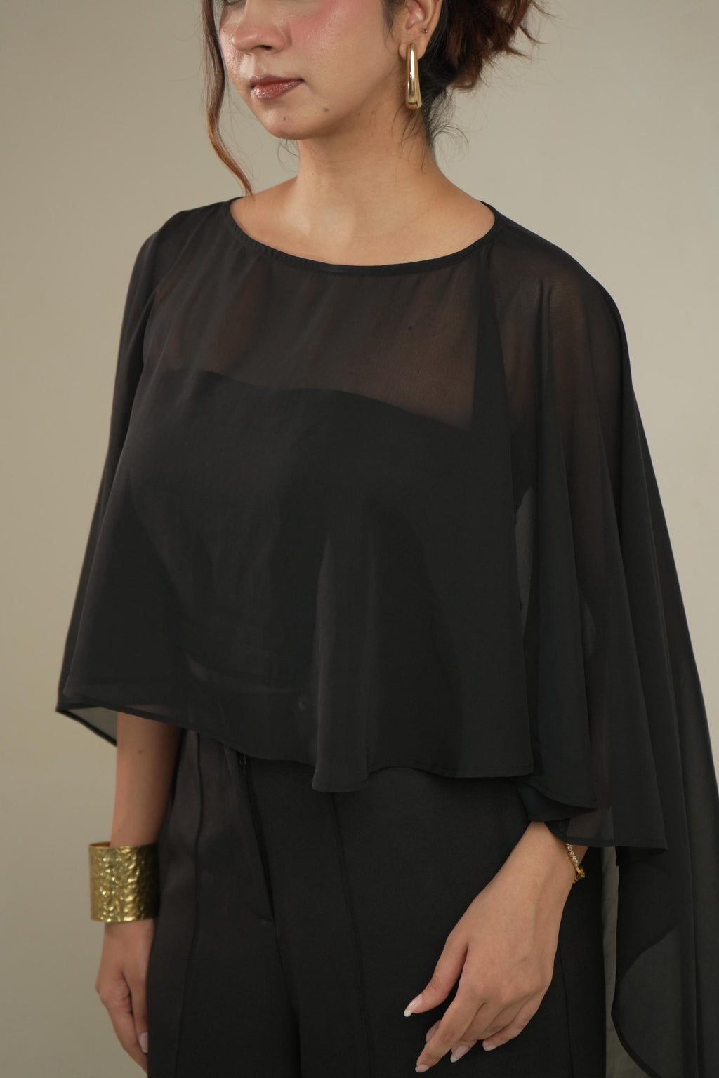 A woman wearing a black sheer draped top with wide flutter sleeves and black pants, photographed against a plain warm beige studio background.