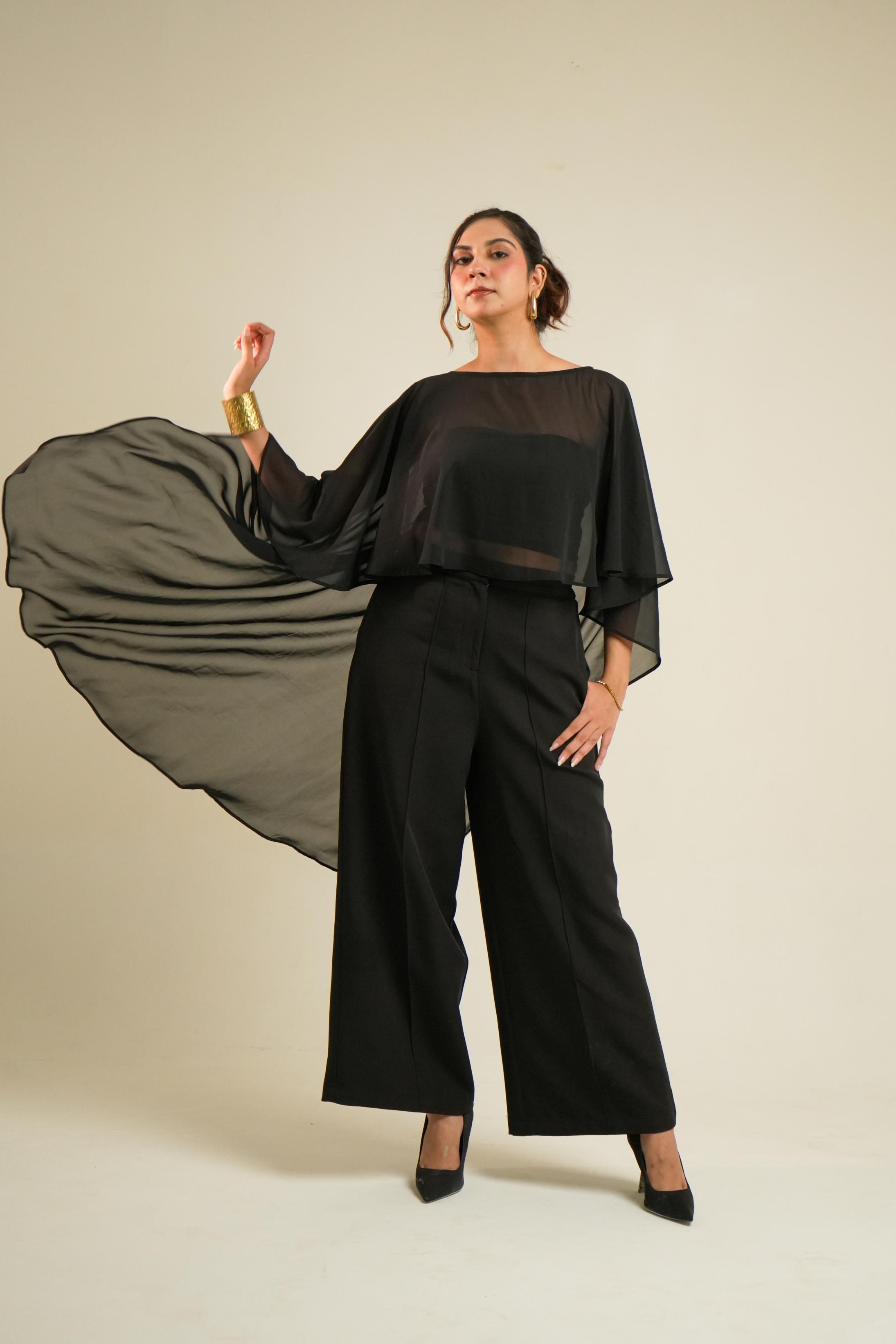 A woman modeling a blacktube topa with cape and black wide-leg trousers, posed against a plain beige studio background.