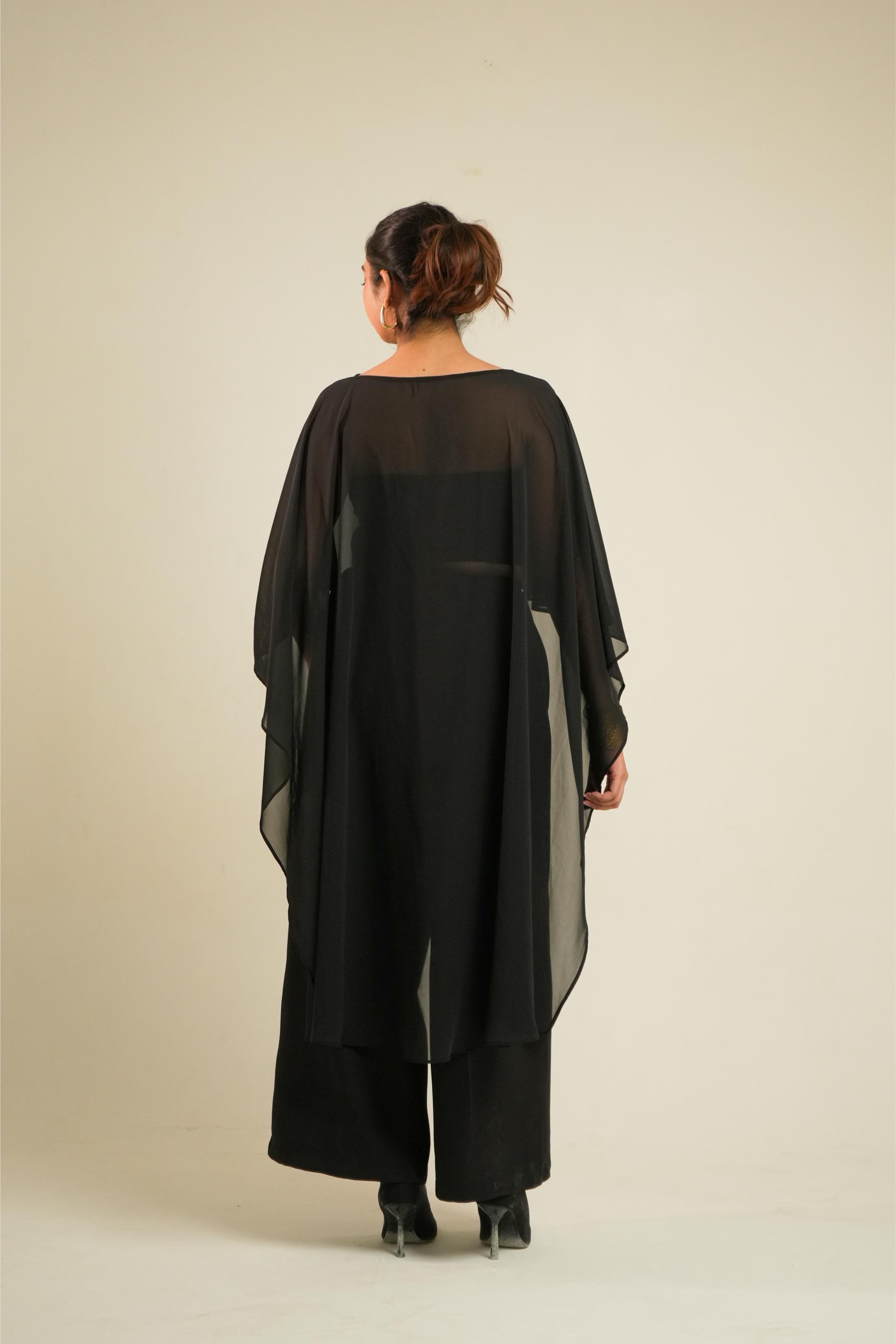 A woman viewed from behind wearing a loose black outfit with a sheer draped overlay and wide-leg pants, standing against a plain light beige studio background.