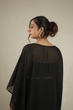 A woman shown from the back wearing a sheer black draped cape-style top or dress against a plain light beige studio background.