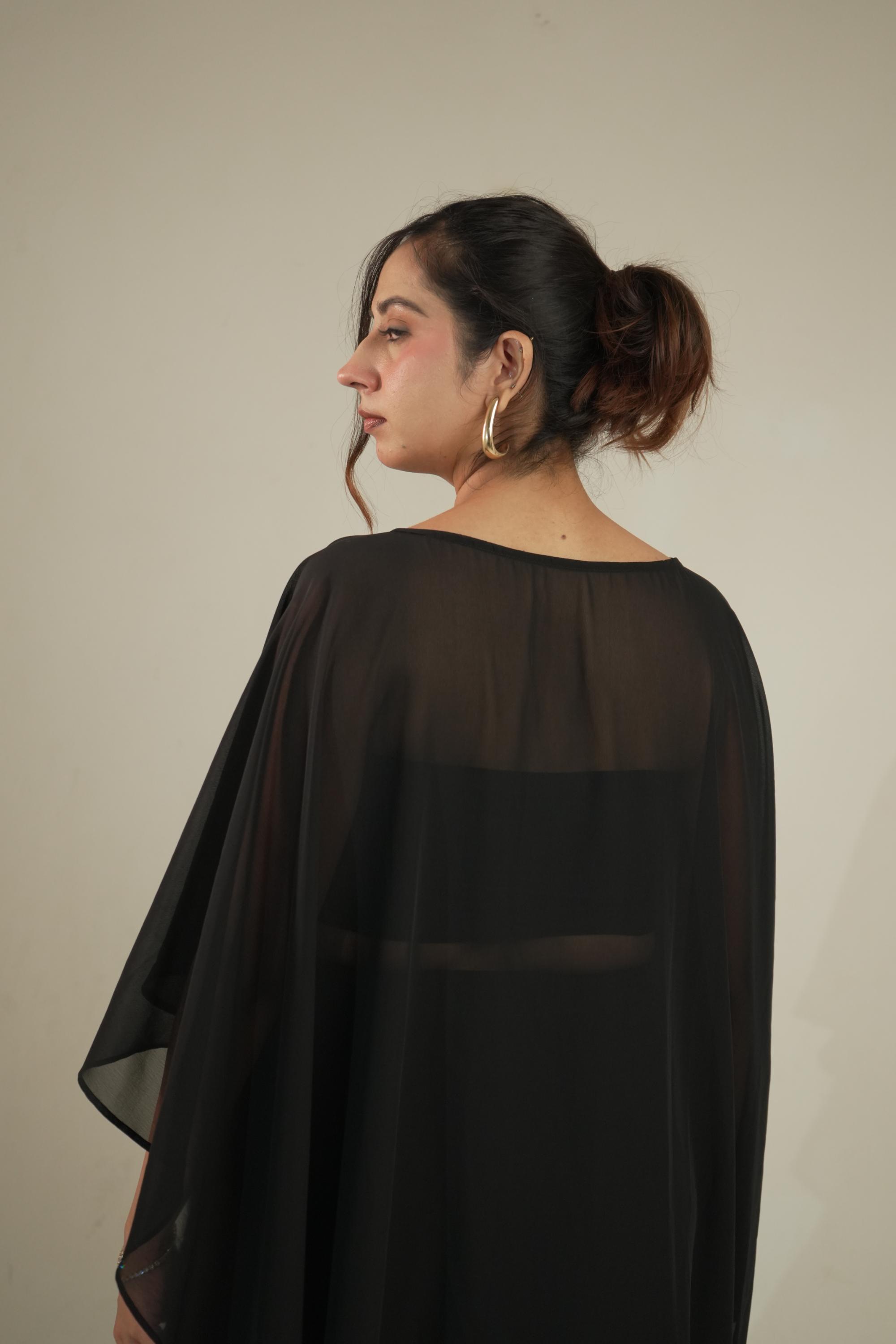 A woman shown from the back wearing a sheer black draped cape-style top or dress against a plain light beige studio background.