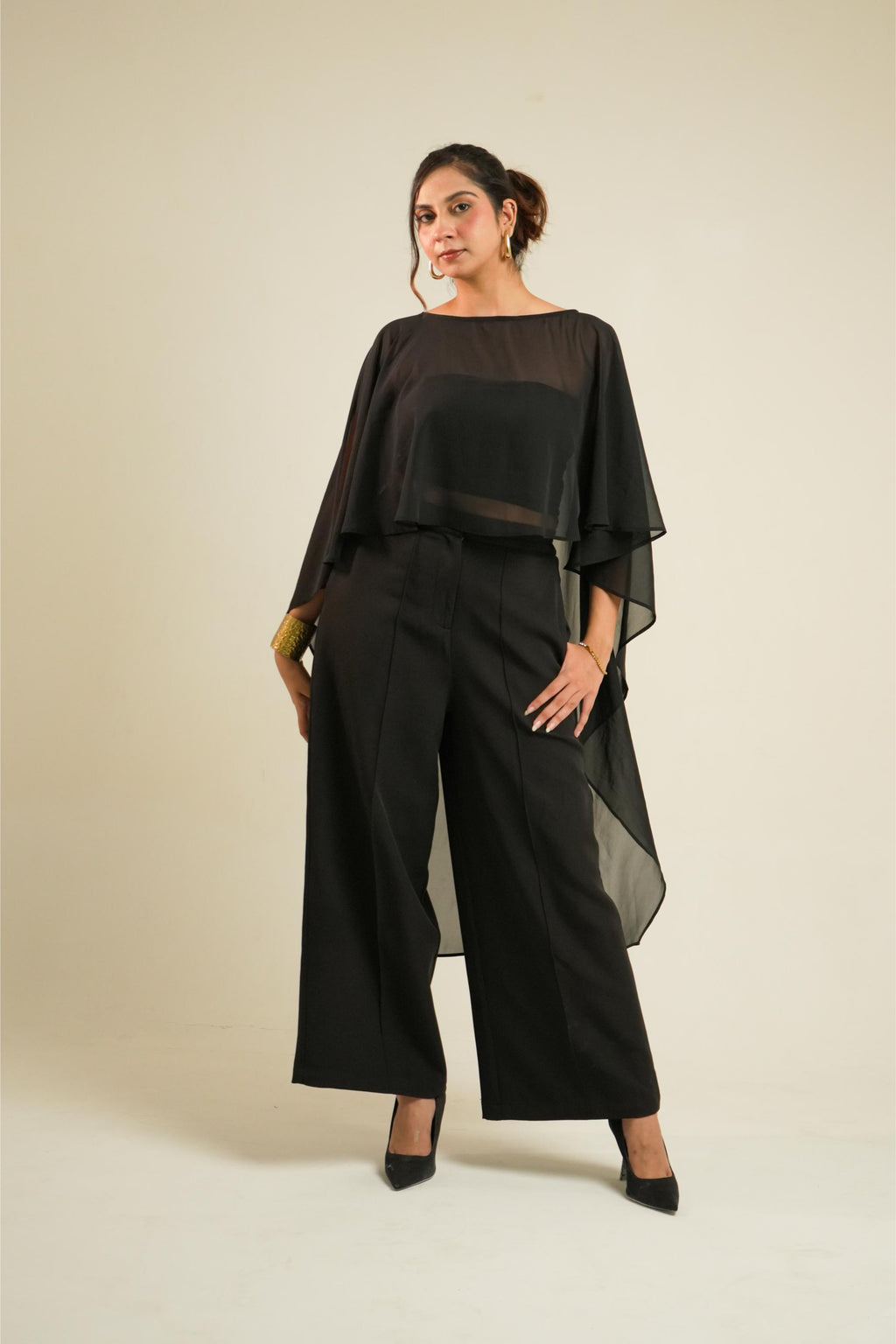 A woman modeling Vrinda Three-Piece Black Co-ord Set | Tube Top, Cape & Wide-Leg Pants standing against a plain beige studio background.