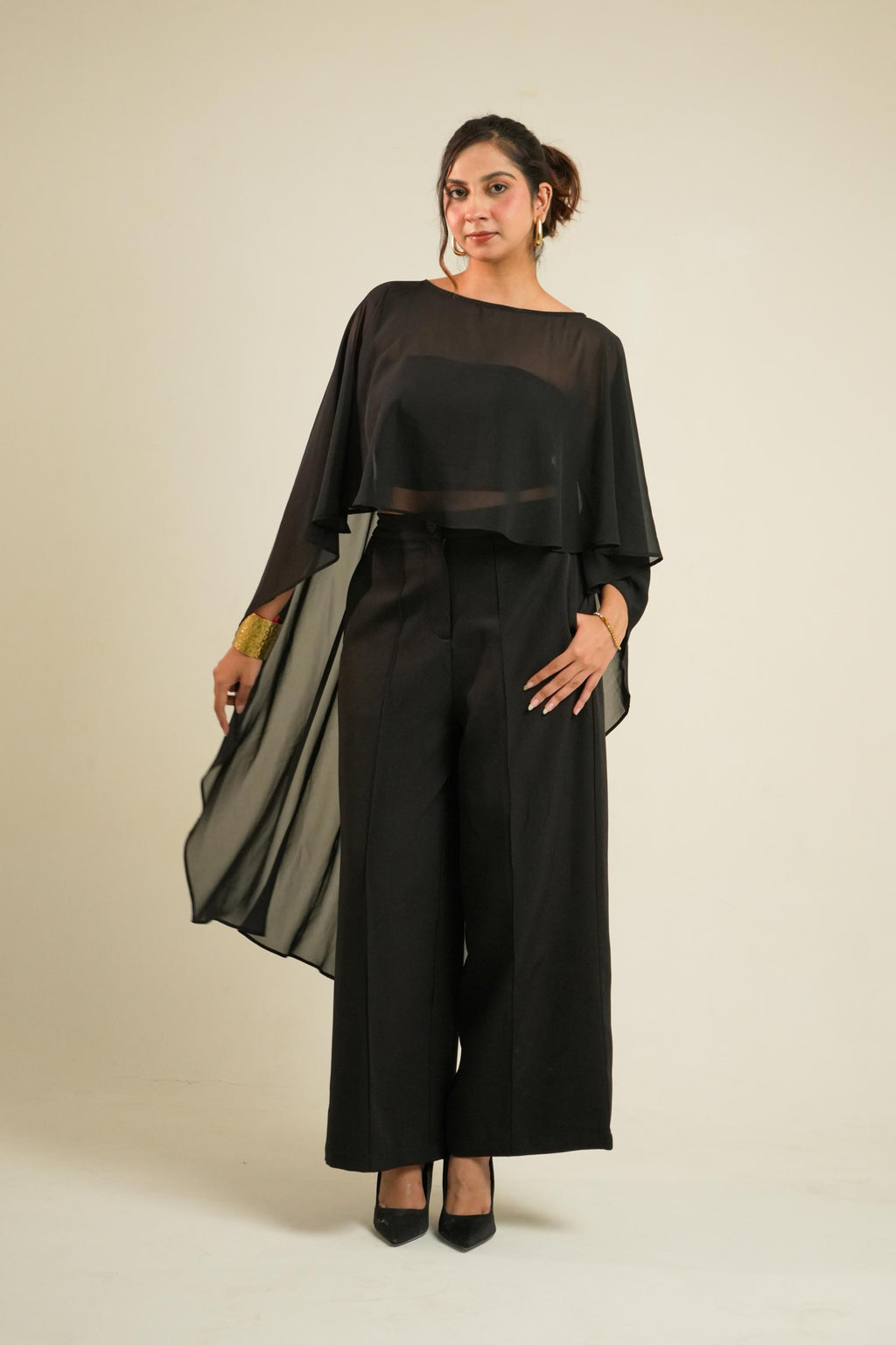 A woman modeling a black two-piece outfit consisting of a sheer draped long-sleeve cape and matching wide-leg trousers, paired with black heels, against a plain beige studio background.