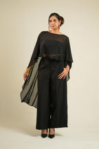A woman modeling a black two-piece outfit consisting of a sheer draped long-sleeve cape and matching wide-leg trousers, paired with black heels, against a plain beige studio background.
