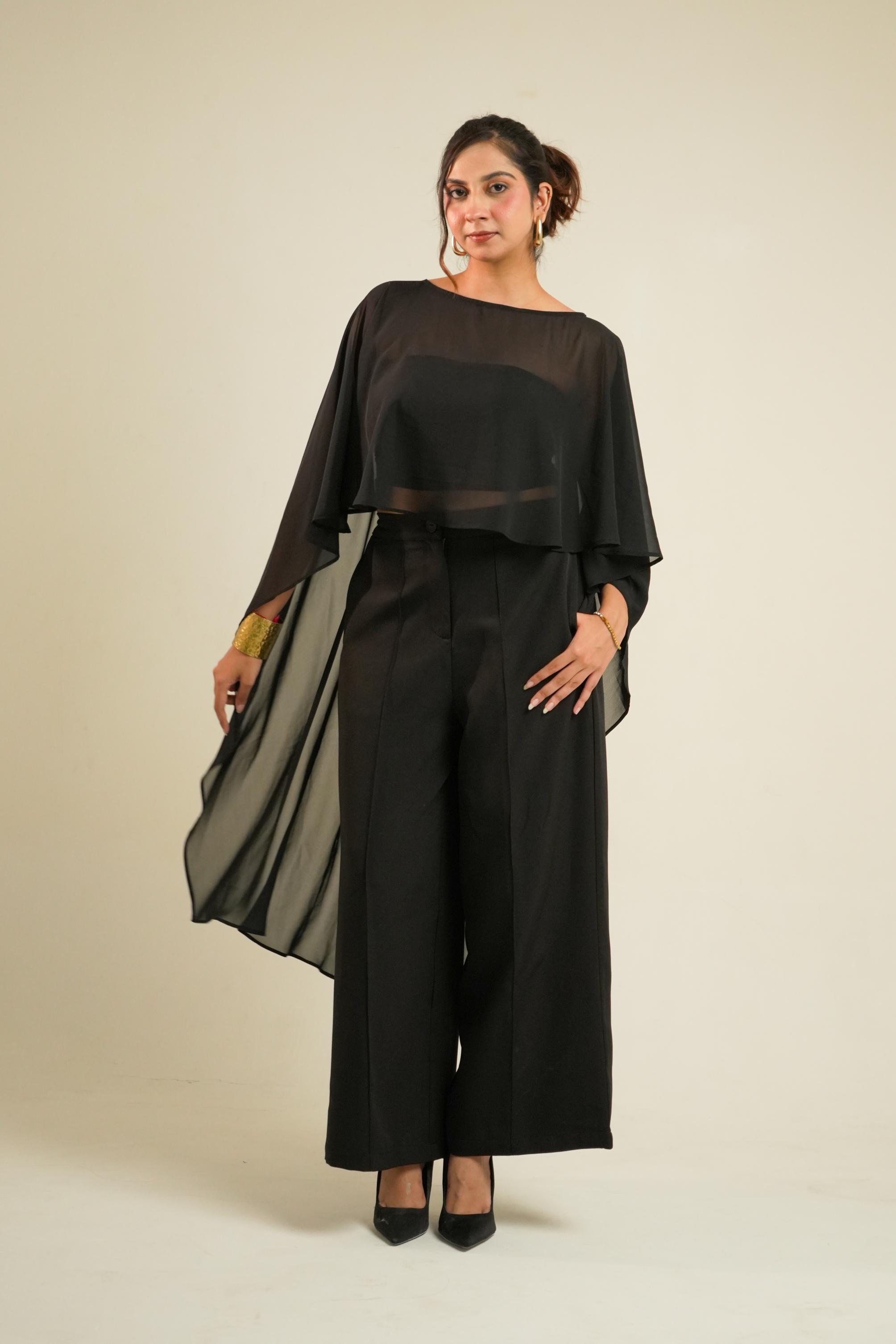 A woman modeling a black two-piece outfit consisting of a sheer draped long-sleeve cape and matching wide-leg trousers, paired with black heels, against a plain beige studio background.