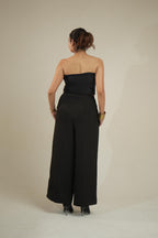 A woman viewed from the back wearing a vrinda black strapless tube top with wide legs, standing against a plain light beige studio background.