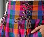 Close-up of a colorful plaid skirt with a lace-up corset-style back, featuring gold eyelets and multicolor stripes in pink, purple, blue, green, orange, and black. A hand is visible holding the skirt.