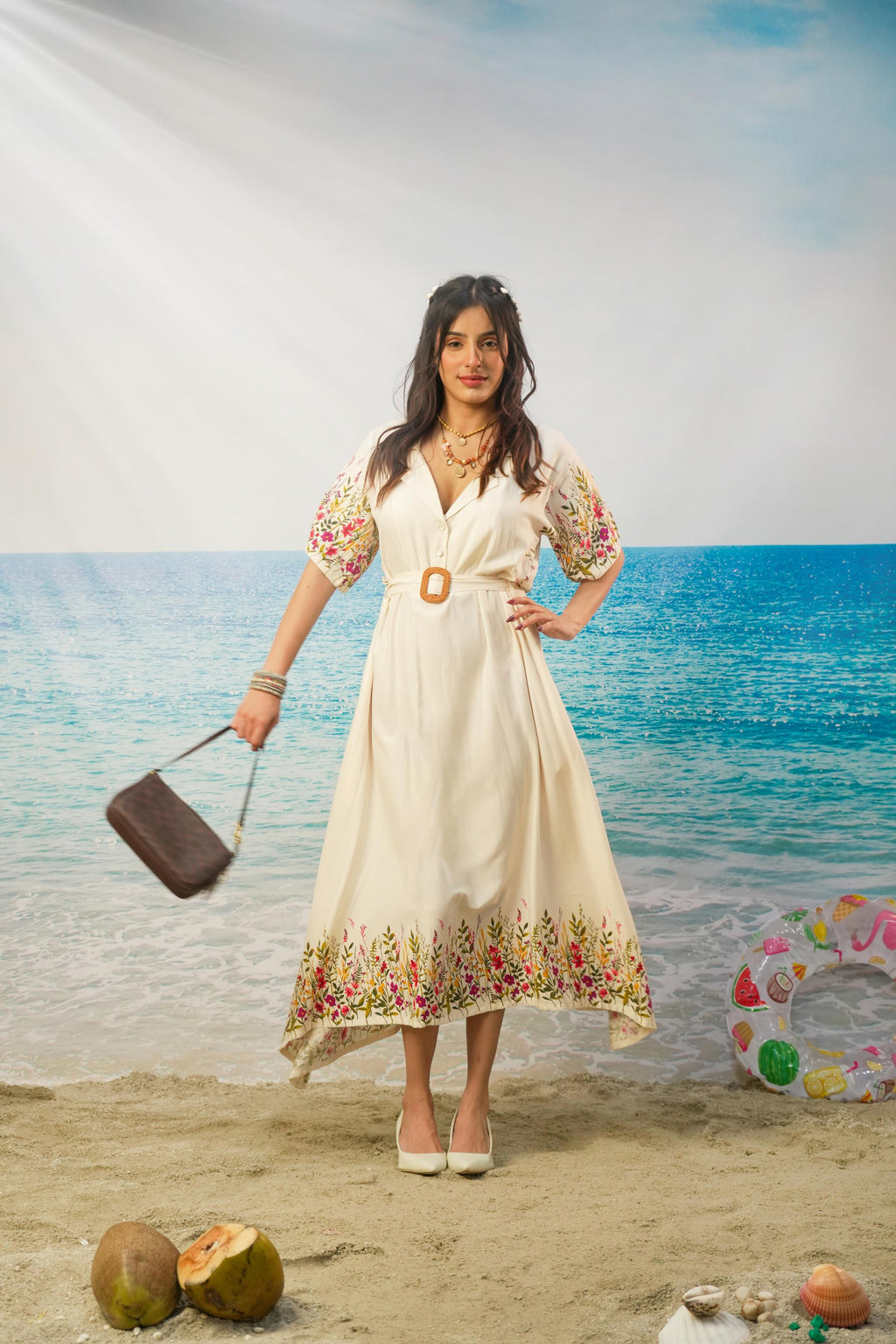 A woman stands on a beach backdrop wearing a cream midi dress with colorful floral print on the sleeves and hem, holding a brown handbag.