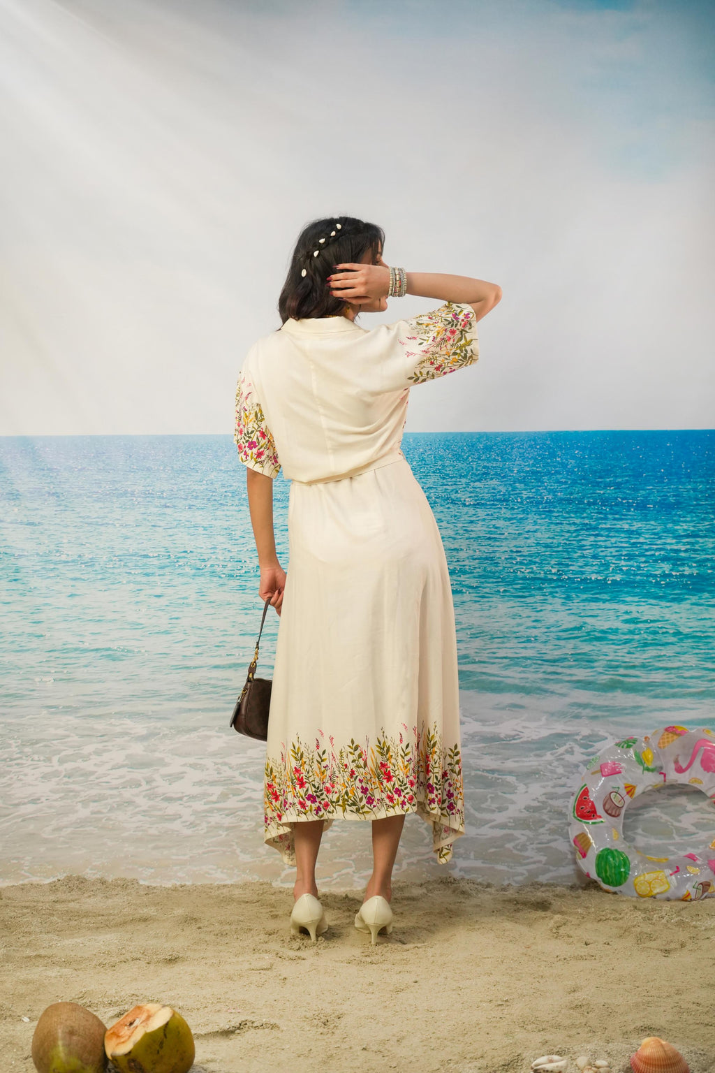 A woman stands on a beach backdrop wearing a cream-colored midi dress with colorful floral print on the sleeves and hem, holding a small black handbag.