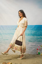 A woman wearing a cream-colored printed midi dress walks on a sandy beach with the ocean in the background, holding a dark brown handbag.