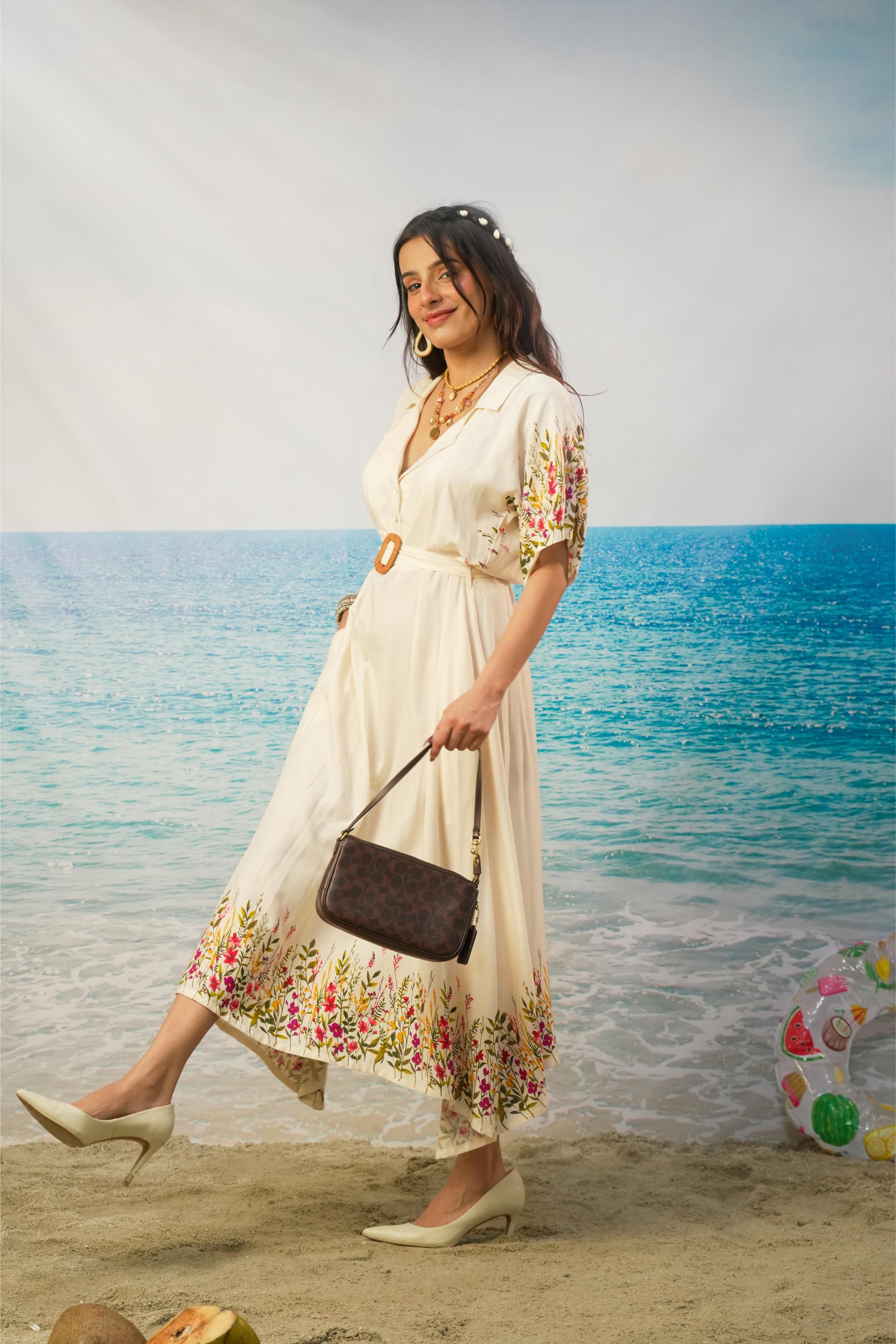 A woman wearing a cream-colored printed midi dress walks on a sandy beach with the ocean in the background, holding a dark brown handbag.