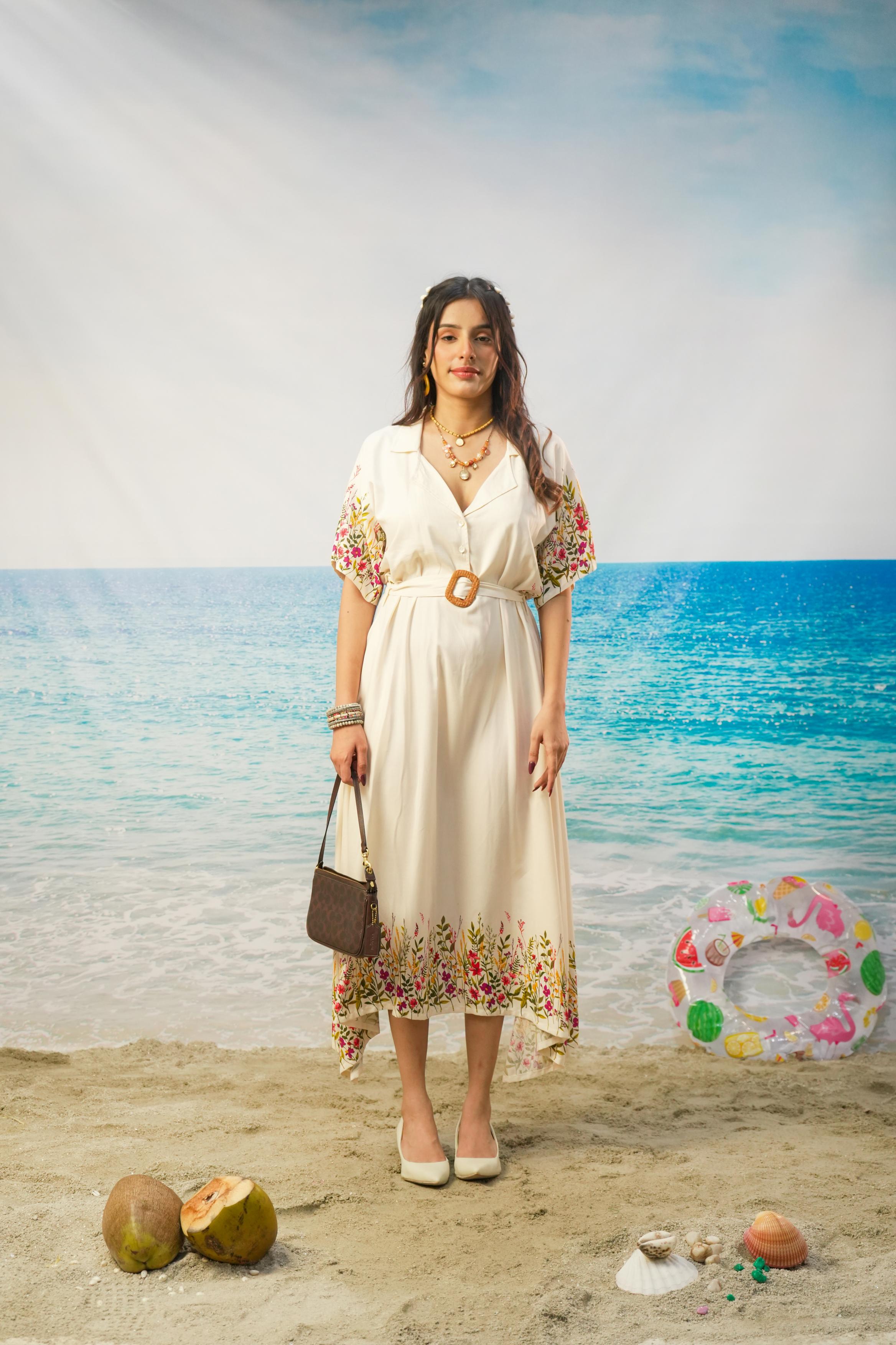 A woman stands on a sandy beach wearing a cream-colored midi dress with colorful floral print on the sleeves and hem, holding a brown handbag. The background shows a blue ocean and sky, with coconuts and a swim ring nearby.