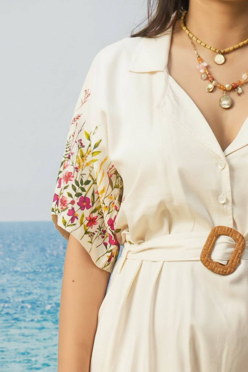 A close-up of a woman wearing a cream-colored collared shirt dress with colorful floral print on the sleeve and a woven belt, set against a bright, overexposed background with a hint of the ocean.