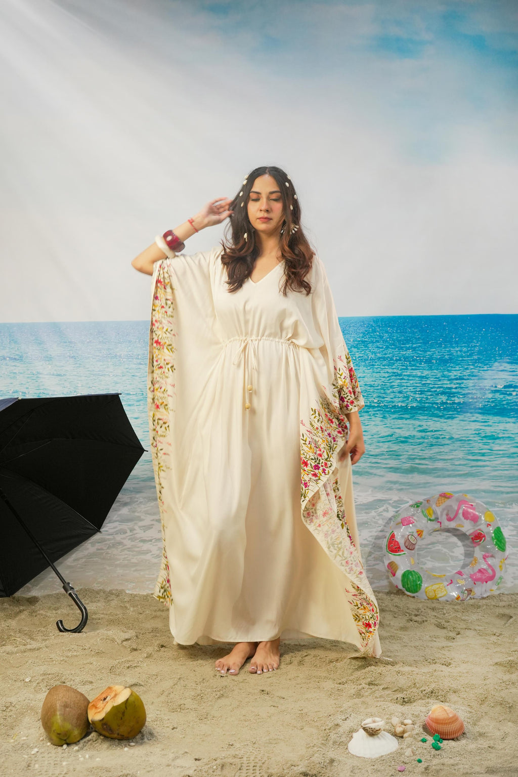 A woman modeling a cream-colored maxi kaftan dress with colorful floral patterns on the borders, standing on a sandy beach backdrop with a blue ocean and sky background.