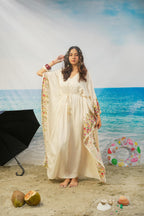 A woman modeling a cream-colored maxi kaftan dress with colorful floral patterns on the borders, standing on a sandy beach backdrop with a blue ocean and sky background.