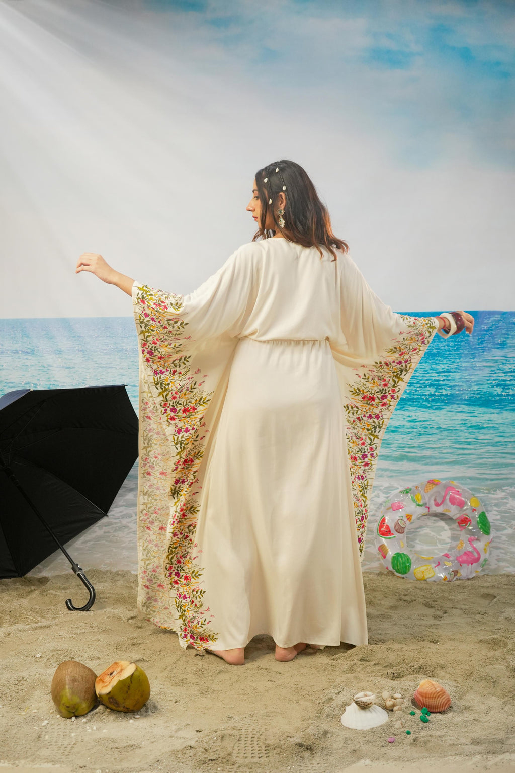 A woman stands on a beach backdrop wearing a flowing cream maxi dress with wide sleeves and colorful floral Printed along the edges. She is photographed from behind, with a beach scene, ocean, sand, umbrella, and inflatable ring around her.
