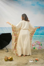 A woman stands on a beach backdrop wearing a flowing cream maxi dress with wide sleeves and colorful floral Printed along the edges. She is photographed from behind, with a beach scene, ocean, sand, umbrella, and inflatable ring around her.