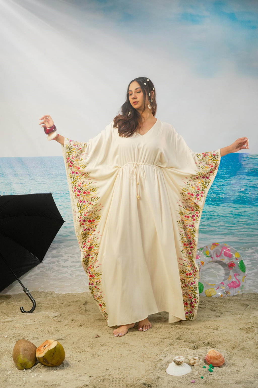 A woman modeling a cream-colored maxi kaftan dress with colorful floral patterns along the wide sleeves, standing on a sandy beach backdrop with a blue ocean and sky background.