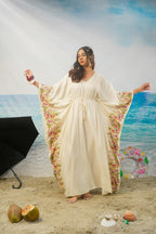A woman modeling a cream-colored maxi kaftan dress with colorful floral patterns along the wide sleeves, standing on a sandy beach backdrop with a blue ocean and sky background.