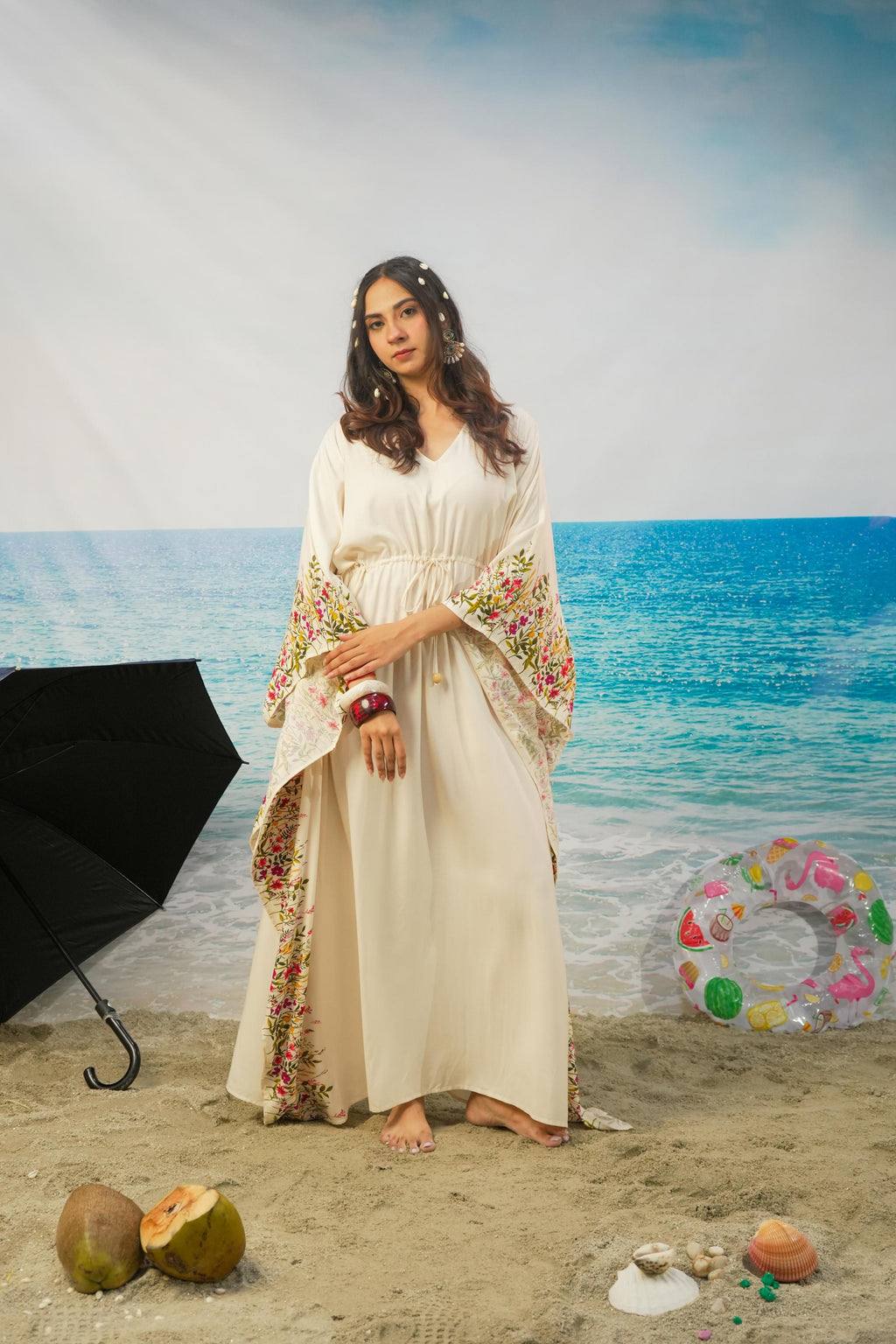 A woman stands barefoot on a sandy beach backdrop wearing a long cream-colored kaftan-style dress with colorful floral printed on the sleeves and hem. A black umbrella, beach coconuts, a pink float, and a beach scene backdrop are visible around her.