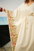 A cream-colored kaftan dress with wide sleeves, featuring a detailed floral and botanical border print in pink, red, and green along the sleeve, shown on a model.
