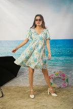 A woman modeling a short-sleeve V-neck Zohra Botanical Cotton Linen Puff Sleeve Dress | Short Resort Dress with a light blue, green, and yellow floral print, standing on a sandy beach backdrop with a blue ocean scene behind her.