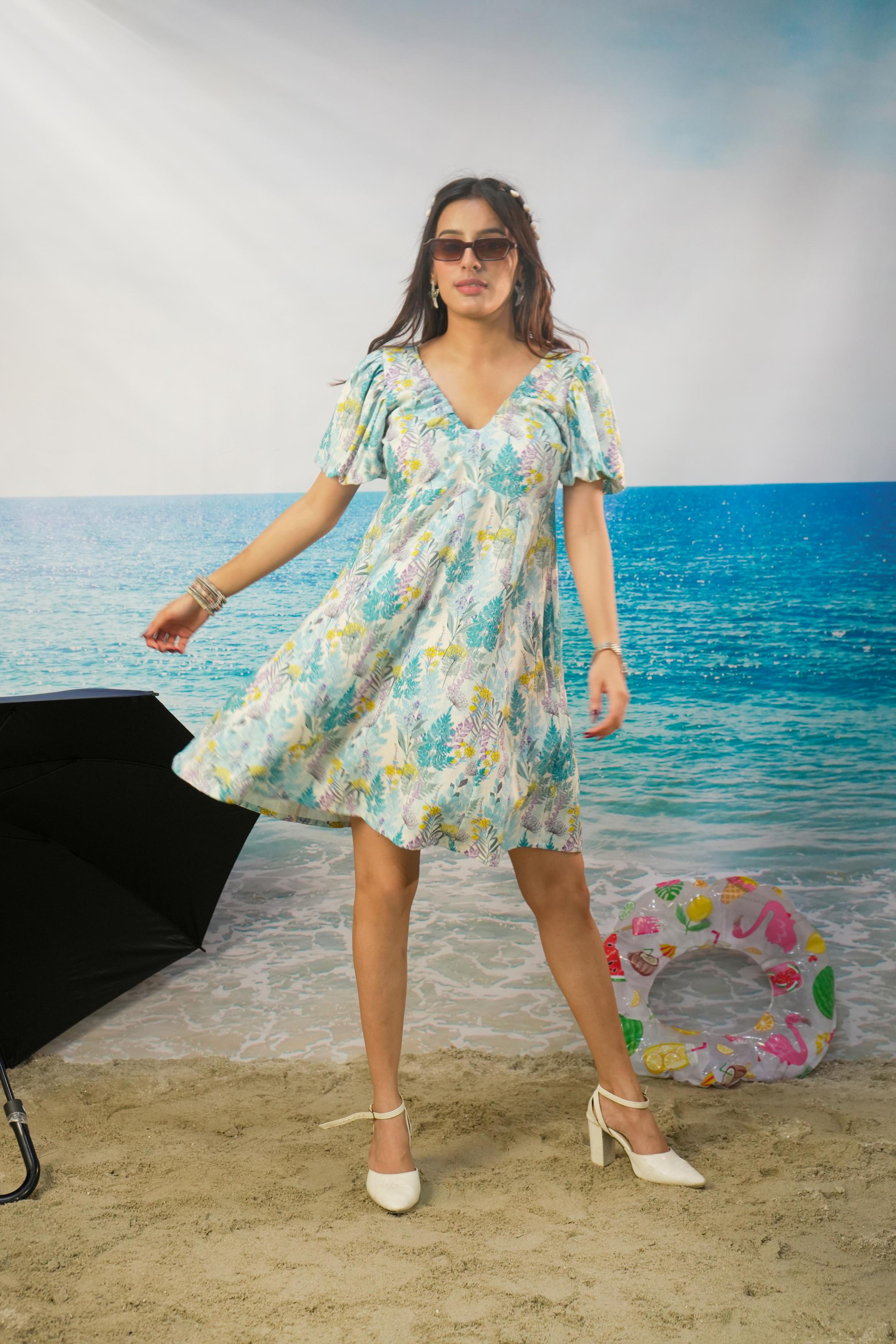 A woman modeling a short-sleeve V-neck Zohra Botanical Cotton Linen Puff Sleeve Dress | Short Resort Dress with a light blue, green, and yellow floral print, standing on a sandy beach backdrop with a blue ocean scene behind her.