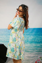 A woman wearing a short-sleeve, off-the-shoulder dress with a tropical floral print in shades of teal, yellow, and purple on a white background, standing in front of a beach backdrop.