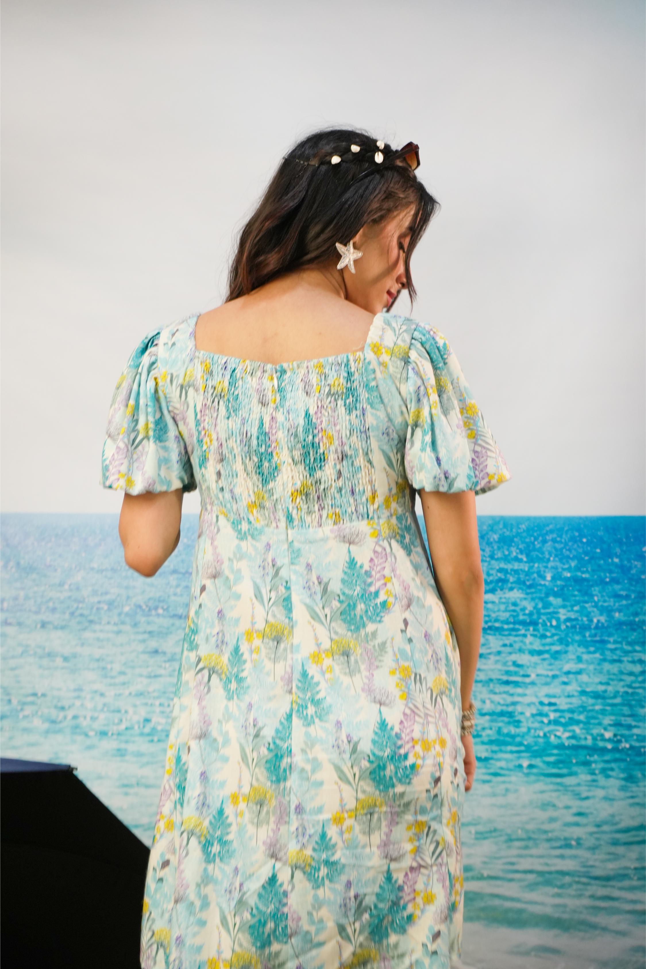A woman wearing a light blue, green, and yellow tropical floral print dress, photographed from behind against a backdrop of a blue ocean and a light gray sky.