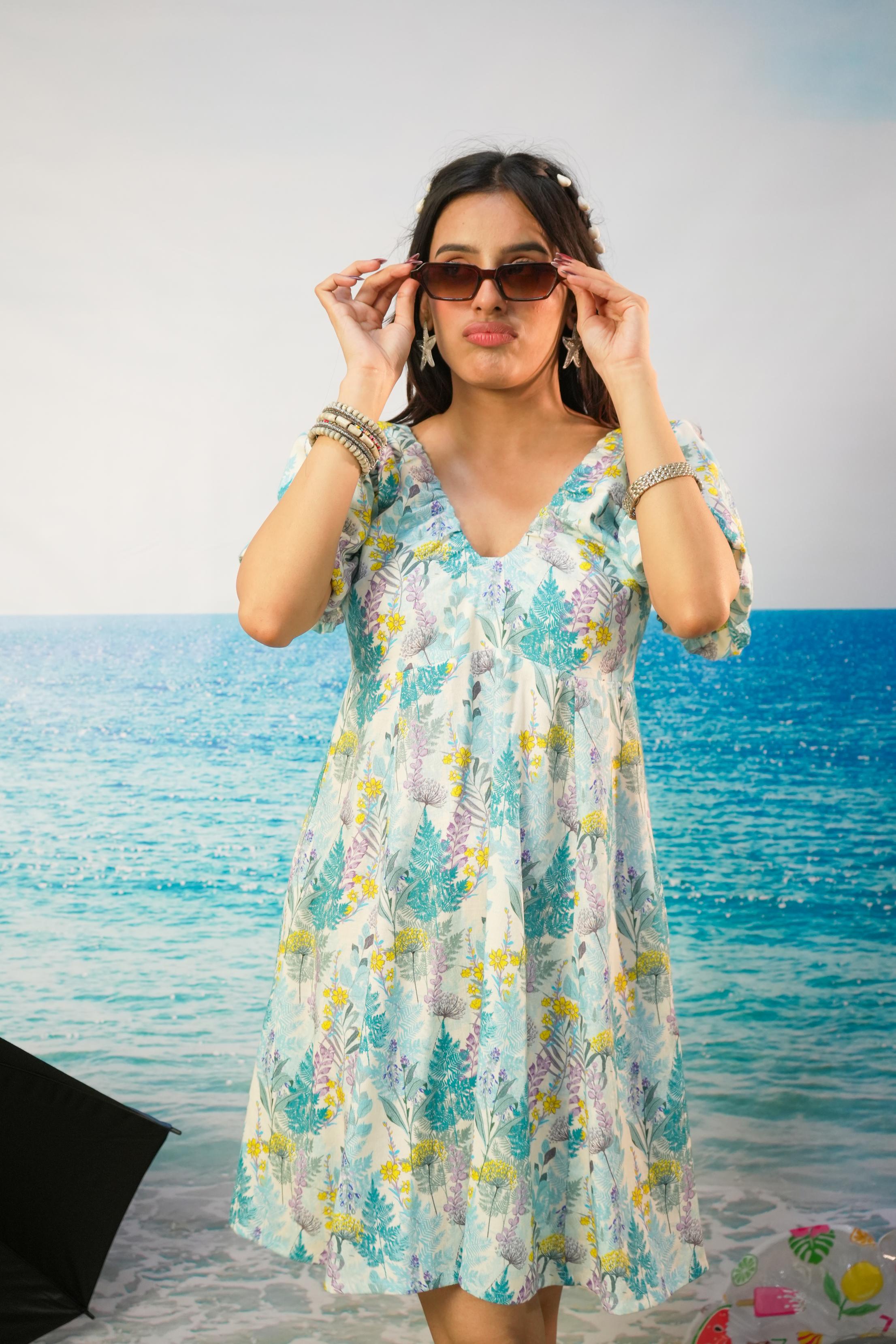 A woman wearing a light blue and white tropical print Zohra Botanical Cotton Linen Puff Sleeve Dress | Short Resort Dress  with puff sleeves, standing in front of a beach backdrop and adjusting her sunglasses.